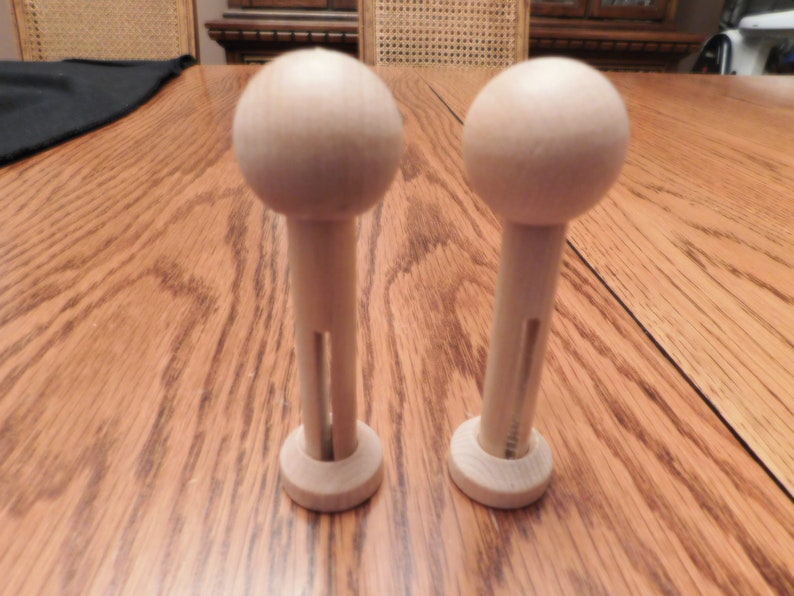 May include: Two unfinished wooden doll pins with a rounded top and a cylindrical stem. The pins have a small circular base and a slot cut into the stem. The items are made of light-colored wood and are displayed on a wooden surface.