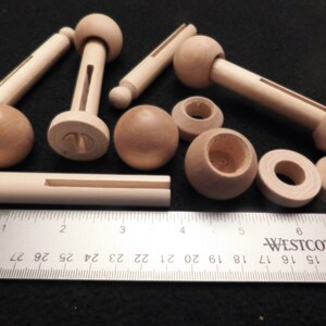 May include: A collection of unfinished wooden craft parts, including dowels, spheres, and turned components. The pieces are light brown and vary in size, with a ruler providing scale in inches. Ideal for woodworking or DIY projects.