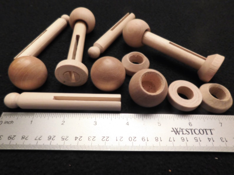 May include: A collection of unfinished wooden craft parts, including dowels, spheres, and turned components. The pieces are light brown and vary in size, with a ruler providing scale in inches. Ideal for woodworking or DIY projects.