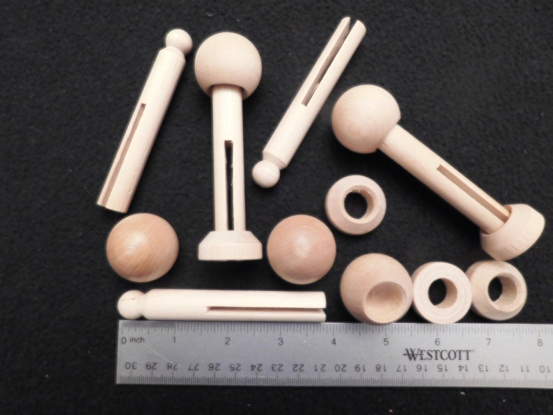 May include: A collection of unfinished wooden craft pieces, including dowels, spheres, and slotted components. A ruler with inch measurements is placed next to the wooden pieces. The items are arranged on a dark surface.
