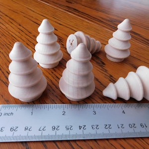 May include: Six unfinished wooden Christmas tree ornaments of varying sizes are arranged on a wooden surface. A ruler is placed near the ornaments to show scale. The trees are light-colored and have a simple, tiered design.
