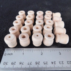 May include: A collection of small, unfinished wooden beads. Each bead has a rounded base and a cylindrical top with a hole. A ruler with inch measurements is in the foreground, showing the size of the beads.