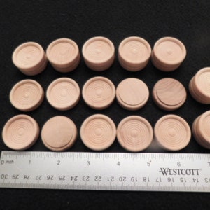 May include: A collection of unfinished wooden game pieces, likely for checkers or a similar board game. The pieces are light brown, circular, and feature concentric circle detailing. A ruler is included for scale.