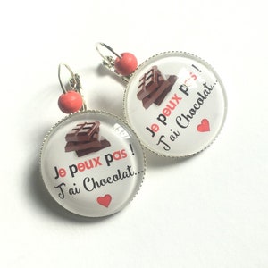 Puede incluir: Un par de pendientes con un dije redondo de vidrio que presenta una barra de chocolate y el texto "Je peux pas! J'ai Chocolat..." en francés. Los dijes son plateados con una cuenta roja en la parte superior del pendiente.