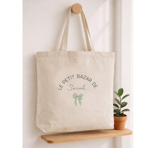 Peut inclure: Sac fourre-tout crème avec le texte "LE PETIT BAZAR DE Sarah" en écriture noire et un motif de nœud vert. Le sac est suspendu à une cheville en bois sur un mur blanc. Une petite plante en pot est posée sur une étagère en bois en dessous.