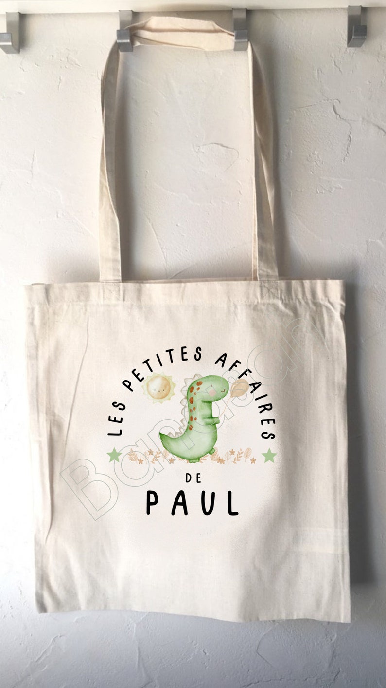 May include: A white canvas tote bag with a green dinosaur illustration and the text "Les Petites Affaires de Paul".