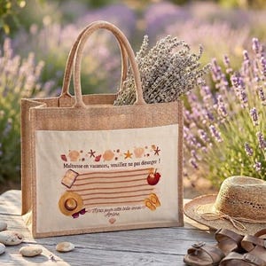 Puede incluir: Un bolso tote beige con un bolsillo delantero de color claro con una ilustraci&oacute;n de tem&aacute;tica playera y texto en franc&eacute;s. La bolsa contiene ramitas de lavanda. Un sombrero de paja y sandalias marrones descansan cerca.