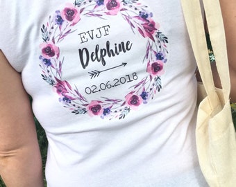May include: White t-shirt with a floral wreath design. The wreath has pink and purple flowers and leaves. The text "EVJF Delphine 02.06.2018" is inside the wreath.