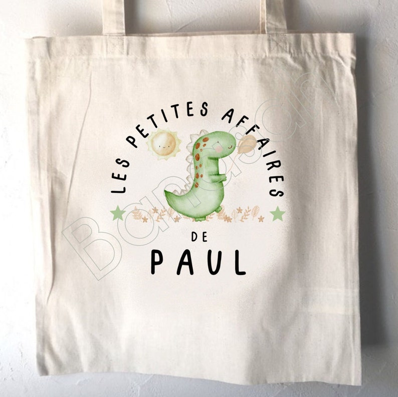 May include: A white canvas tote bag with a green dinosaur illustration and the text "Les Petites Affaires de Paul".