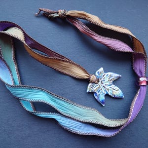 May include: A multi-coloured ribbon necklace with a ceramic leaf pendant. The ribbon is brown, purple, and blue. The leaf is blue, green, and white.