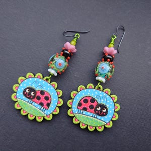 May include: A pair of dangle earrings featuring a ladybug design with black and red spots. The earrings are made with colorful beads and have a green and blue background.