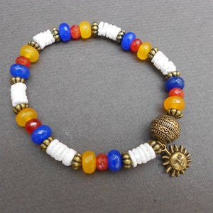 Op de afbeelding: Een kleurrijke kralen armband met witte, gele, rode en blauwe kralen. De armband heeft een gouden zon hanger en een ronde kraal met een geometrisch patroon.