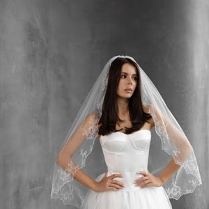 K&ouml;nnte beinhalten: Ein wei&szlig;es Brautkleid mit tr&auml;gerlosem Oberteil und einem vollen T&uuml;llrock. Die Braut tr&auml;gt einen durchsichtigen Schleier mit floralen Spitzendetails an den R&auml;ndern. Der Hintergrund ist einfarbig grau.
