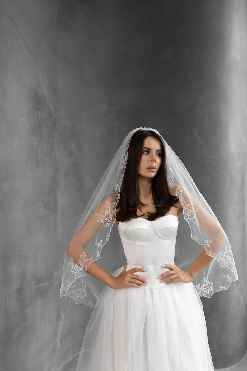 K&ouml;nnte beinhalten: Ein wei&szlig;es Brautkleid mit tr&auml;gerlosem Oberteil und einem vollen T&uuml;llrock. Die Braut tr&auml;gt einen durchsichtigen Schleier mit floralen Spitzendetails an den R&auml;ndern. Der Hintergrund ist einfarbig grau.