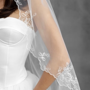 K&ouml;nnte beinhalten: Ein durchsichtiger wei&szlig;er Brautschleier mit zarter floraler Spitzenborte. Der Schleier ist &uuml;ber einem wei&szlig;en Brautkleid drapiert und zeigt die aufwendigen Stickereidetails. Das Blumenmuster umfasst Rosen und Bl&auml;tter.