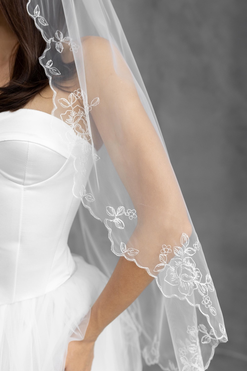 K&ouml;nnte beinhalten: Ein durchsichtiger wei&szlig;er Brautschleier mit zarter floraler Spitzenborte. Der Schleier ist &uuml;ber einem wei&szlig;en Brautkleid drapiert und zeigt die aufwendigen Stickereidetails. Das Blumenmuster umfasst Rosen und Bl&auml;tter.