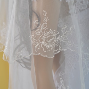 K&ouml;nnte beinhalten: Ein wei&szlig;er Brautschleier mit zarten floralen Stickereien. Der Schleier hat einen gewellten Rand und aufwendige Spitzendetails, die einen eleganten und romantischen Look erzeugen. Das Design umfasst gestickte Rosen und Bl&auml;tter, perfekt f&uuml;r eine Hochzeit.