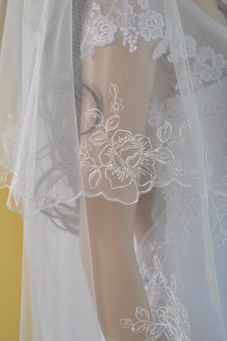 K&ouml;nnte beinhalten: Ein wei&szlig;er Brautschleier mit zarten floralen Stickereien. Der Schleier hat einen gewellten Rand und aufwendige Spitzendetails, die einen eleganten und romantischen Look erzeugen. Das Design umfasst gestickte Rosen und Bl&auml;tter, perfekt f&uuml;r eine Hochzeit.