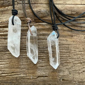 May include: Three clear quartz crystal pendants on black leather cords. The crystals vary in shape and size, each with a unique, natural form. The pendants are displayed against a rustic, weathered wooden background.