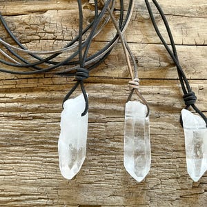 Puede incluir: Tres colgantes de cristal de cuarzo transparente con cordones de cuero. Los cristales varían en tamaño y forma, cada uno suspendido de un cordón de cuero anudado. Los cordones son negros y marrones, y se muestran sobre un fondo de madera envejecida.
