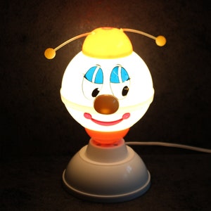 May include: A vintage novelty lamp shaped like a cartoon face with a yellow hat and antennae. The lamp has a white globe with blue eyes, a brown nose, and a red smiling mouth. The base is white and orange.
