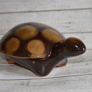 Può includere: Un portagioie in ceramica a forma di tartaruga con una lucida smaltatura marrone scuro. Il guscio presenta macchie circolari color beige chiaro, mentre la testa, le zampe e la parte inferiore sono marrone scuro. La base rivela l'argilla arancione.