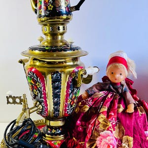 May include: A vintage brass samovar with a painted floral design, a black handle, and a matching tray. A doll in a traditional dress is next to the samovar. The samovar is a traditional Russian tea kettle.