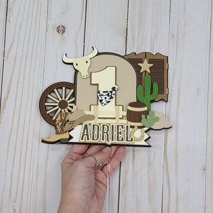 May include: A brown and tan cake topper with a cowboy theme. It features a number one, a cowboy hat, a boot, a wagon wheel, a cactus, a barrel, a bull's head, a saloon door, and a horseshoe. The name "Adriel" is written on a banner.