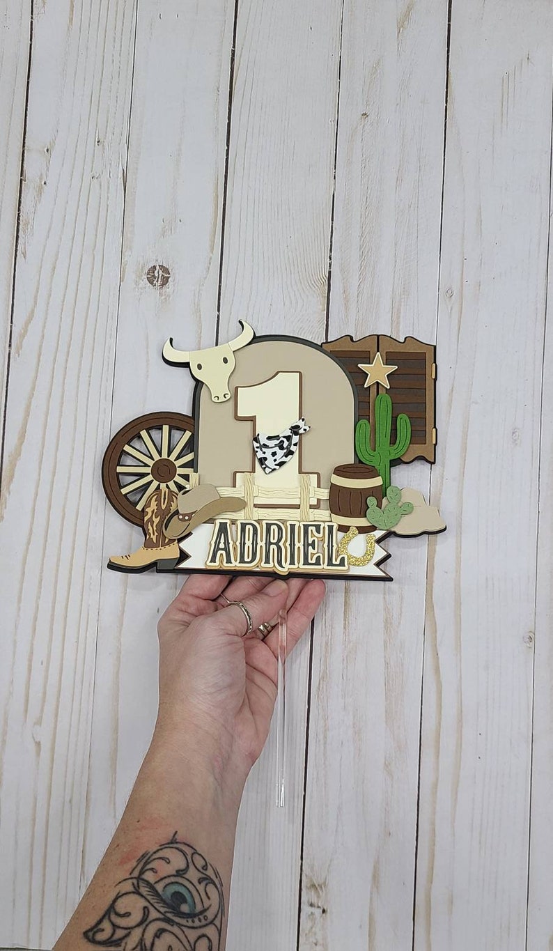 May include: A brown and tan cake topper with a cowboy theme. It features a number one, a cowboy hat, a boot, a wagon wheel, a cactus, a barrel, a bull's head, a saloon door, and a horseshoe. The name "Adriel" is written on a banner.