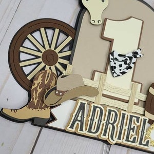 May include: A brown, white, and black cowboy-themed cake topper with a wagon wheel, a cowboy hat, a boot, a bull, and the number one. The name "Adriel" is written in black letters with a white outline.