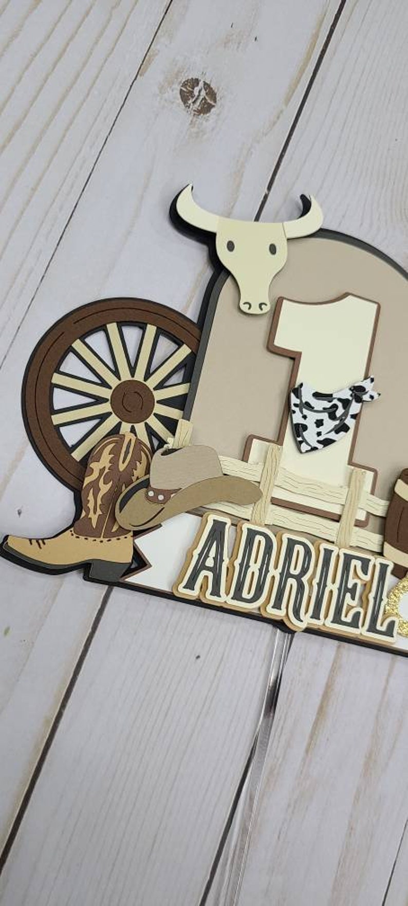 May include: A brown, white, and black cowboy-themed cake topper with a wagon wheel, a cowboy hat, a boot, a bull, and the number one. The name "Adriel" is written in black letters with a white outline.