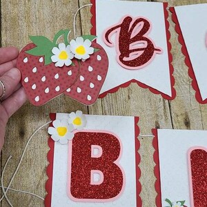 May include: A red and white banner with the letters "Em" and "B" decorated with red glitter and strawberries. The banner is strung together with white string.