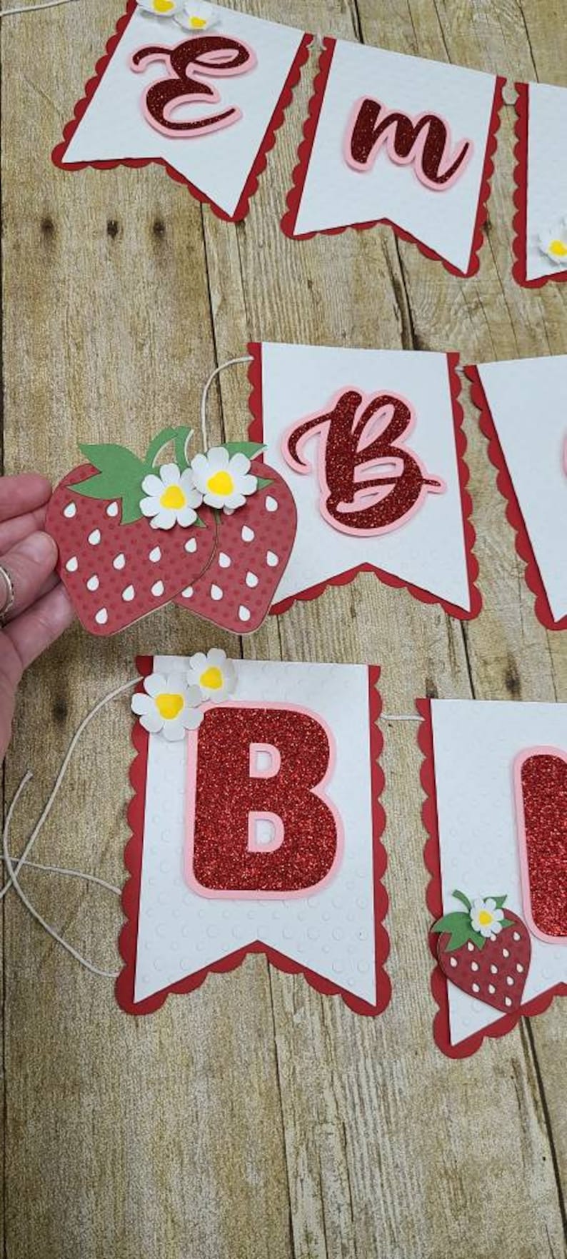 May include: A red and white banner with the letters "Em" and "B" decorated with red glitter and strawberries. The banner is strung together with white string.