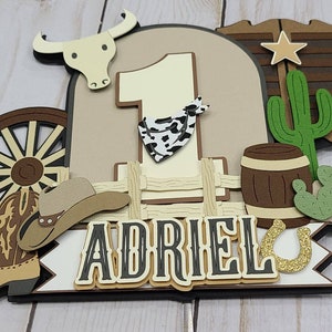 May include: A brown, tan, and green cowboy-themed cake topper with a number one, a cowboy hat, boots, a wagon wheel, a cactus, a barrel, and a horseshoe. The name "Adriel" is written on the topper.