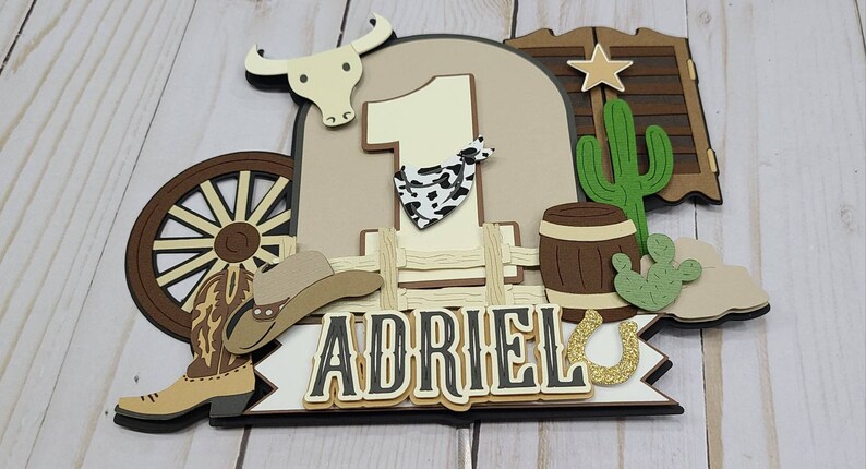May include: A brown, tan, and green cowboy-themed cake topper with a number one, a cowboy hat, boots, a wagon wheel, a cactus, a barrel, and a horseshoe. The name "Adriel" is written on the topper.