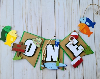 May include: A colorful paper banner with the word "ONE" in white letters, decorated with fishing-themed elements like fish, a fishing rod, a fishing lure, and a "No Fishing" sign. The banner is perfect for a first birthday party.
