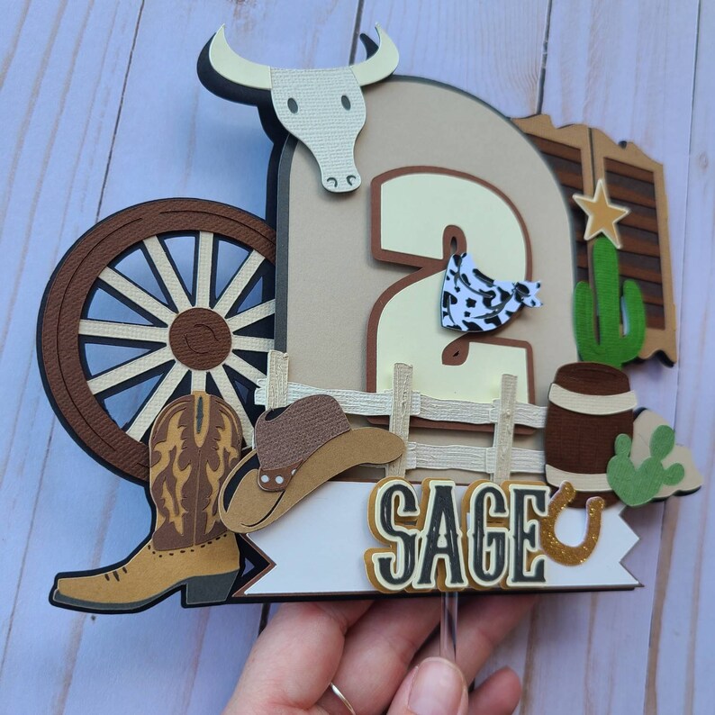 May include: A brown, white, and yellow cowboy-themed cake topper with a number 2, a wagon wheel, a cowboy hat, a cowboy boot, a cactus, a barrel, and a horseshoe. The name "SAGE" is written on a white banner.