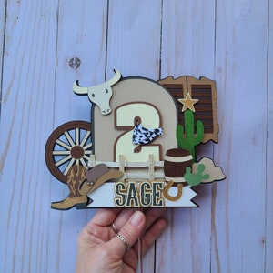May include: A brown and tan cowboy themed cake topper with a number 2, a cowboy hat, a wagon wheel, a cactus, a horseshoe, and the name "Sage".