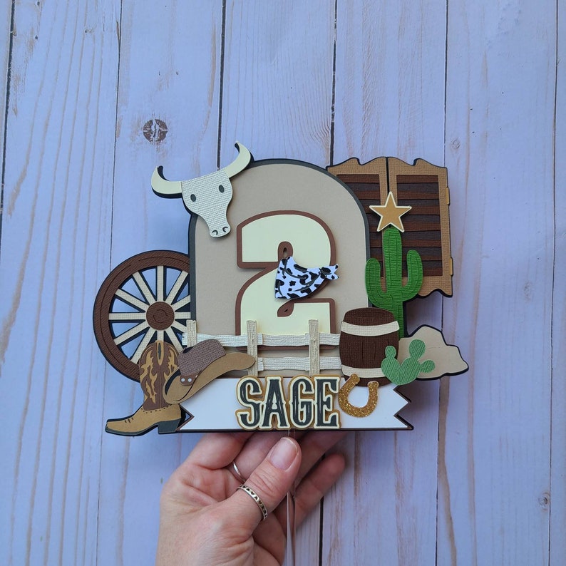 May include: A brown and tan cowboy themed cake topper with a number 2, a cowboy hat, a wagon wheel, a cactus, a horseshoe, and the name "Sage".