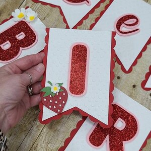 May include: A red and white banner with the word "Birthday" spelled out in red glitter letters. Each letter is on a separate white card with a red scalloped border. Each card also has a red glitter strawberry with a white flower on it.