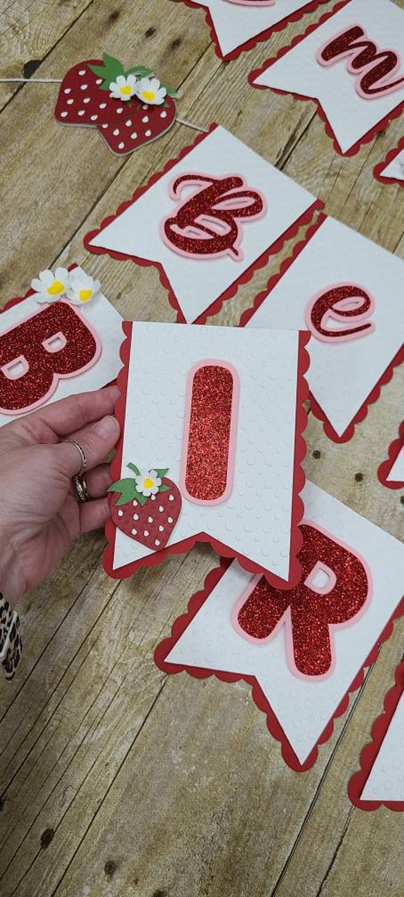 May include: A red and white banner with the word "Birthday" spelled out in red glitter letters. Each letter is on a separate white card with a red scalloped border. Each card also has a red glitter strawberry with a white flower on it.