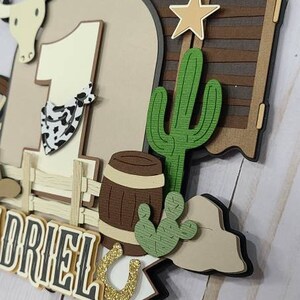 May include: A brown and white cowboy-themed cake topper with a number one, a cactus, a barrel, a horseshoe, and the name "DRIEL".