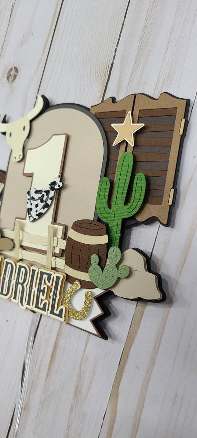 May include: A brown and white cowboy-themed cake topper with a number one, a cactus, a barrel, a horseshoe, and the name "DRIEL".