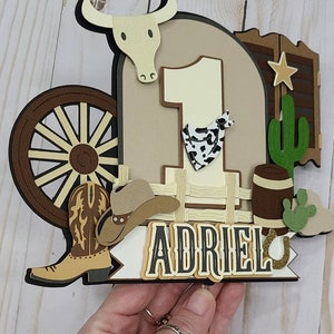 May include: A brown and tan cowboy-themed cake topper with a large number one, a cowboy hat, a boot, a wagon wheel, a cactus, a barrel, and the name "Adriel".