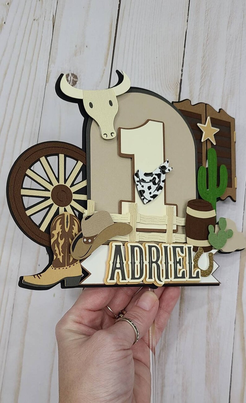 May include: A brown and tan cowboy-themed cake topper with a large number one, a cowboy hat, a boot, a wagon wheel, a cactus, a barrel, and the name "Adriel".
