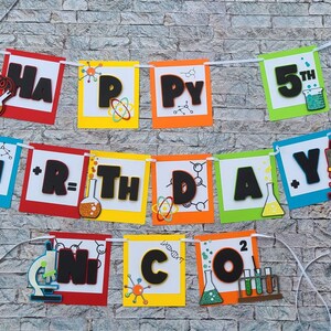 May include: A colorful banner with the words "HAPPY 5TH BIRTHDAY" in black letters. The banner features science-themed decorations, including beakers, test tubes, and a microscope. The banner is strung on a white string.