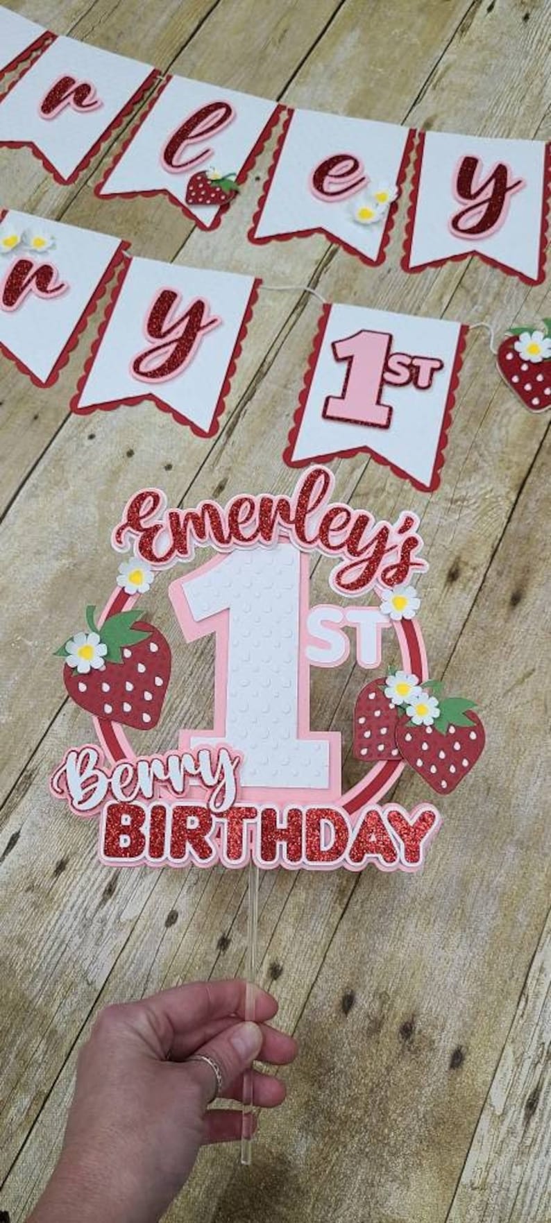 May include: A pink and red cake topper with the text "Emerley's 1st Berry Birthday" and a large white number one with red polka dots. The topper is decorated with red strawberries and white flowers.
