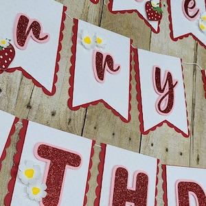 May include: A white banner with red scalloped edges and red glitter letters that spell out "Happy Birthday". Each letter is outlined in pink and has a small strawberry or flower embellishment.