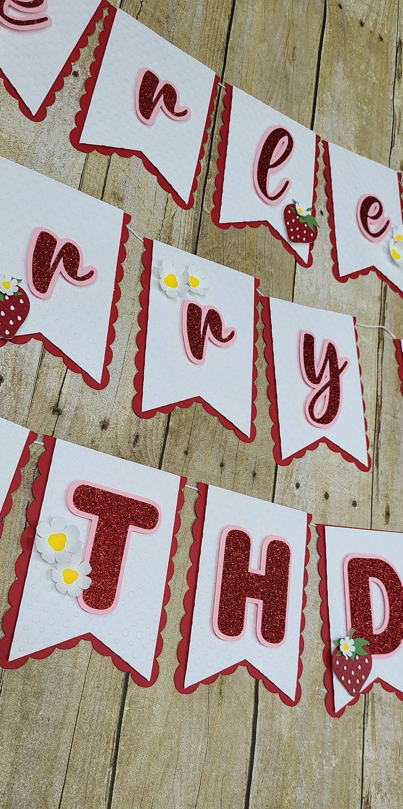 May include: A white banner with red scalloped edges and red glitter letters that spell out "Happy Birthday". Each letter is outlined in pink and has a small strawberry or flower embellishment.