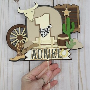 May include: A brown, white, and gold cowboy-themed cake topper with the number 1, a cowboy boot, a cowboy hat, a wagon wheel, a cactus, a barrel, and a horseshoe. The name "ADRIEL" is written on the topper.